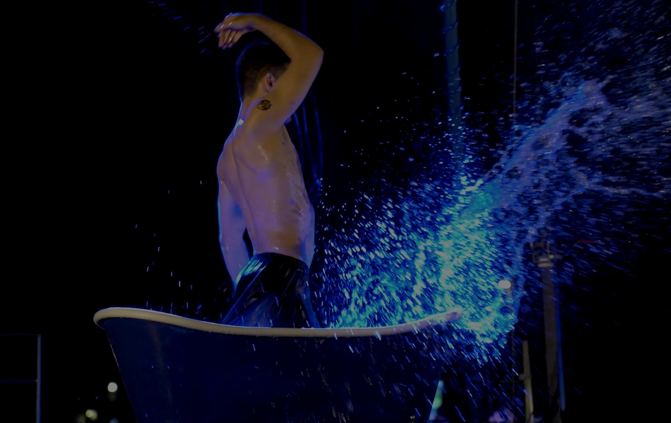 Acróbata sobre el escenario dentro de una bañera, levantando una ola de agua iluminada en azul